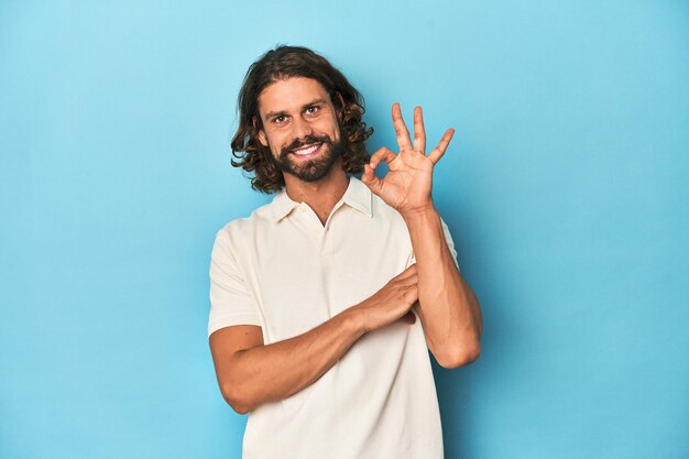 Photo longhaired man in a white polo blue studio winks an eye and holds an okay gesture with hand