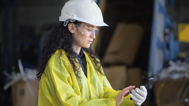 안전모를 쓴 긴 머리 관리자는 폐기물 공장에서 작업 과정을 보고하기 위해 스마트폰에서 연락처를 검색합니다. 감독자는 폐기물 분류 공장을 제어합니다.
