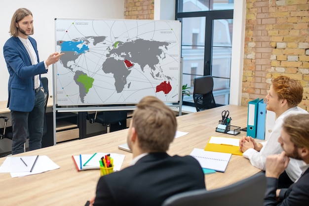 Longhaired smart man talking standing near stand and sitting concerned colleagues in office