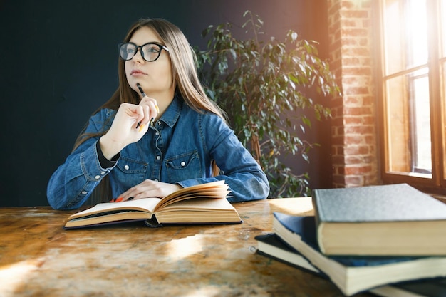 Longhaired student girl wearing glasses making notes from book while preparing for test or exam