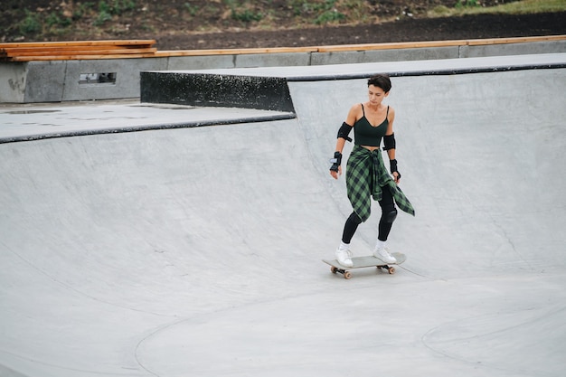 쿼터 파이프 서클에서 보드를 타고 있는 여성 스케이팅 선수의 롱샷. 그녀는 흰색 운동화와 함께 어두운 옷을 입고 있습니다.