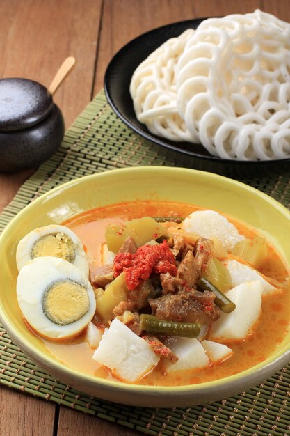 Lontong Sayur Padang, Vegetable Curry with Rice Pressed Cake, Served with Boiled Egg. Served on Wooden Table with Yellow Plate. Served with Krupuk Warung on Background