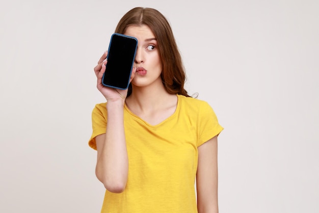 Смотри, рекламируй здесь! Портрет забавной женщины молодого возраста в футболке, закрывающей половину лица телефона с пустым экраном, с задумчивым сомневающимся выражением лица. Крытая студия снята на сером фоне.
