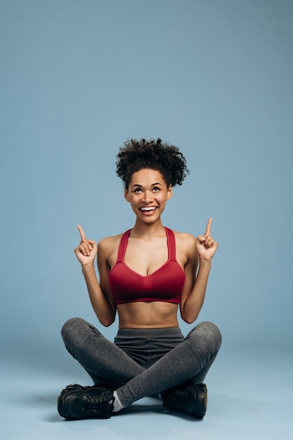 Look at advertise. Portrait of pleasantly surprised sportswoman pointing up and looking with happy face, while sitting at the lotus pose. Indoor studio shoot isolated