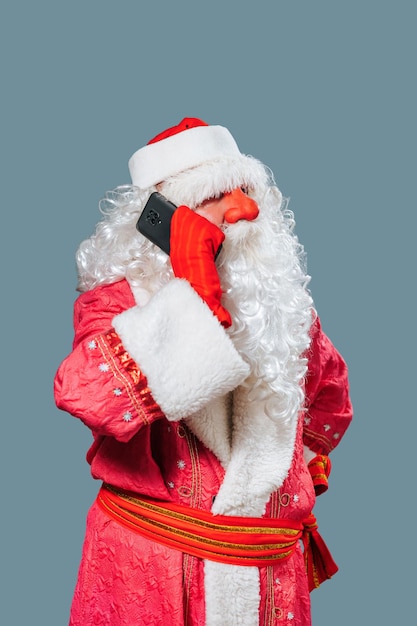 Look at camera blue background close up front view portrait Santa Claus speaking on the smartphone