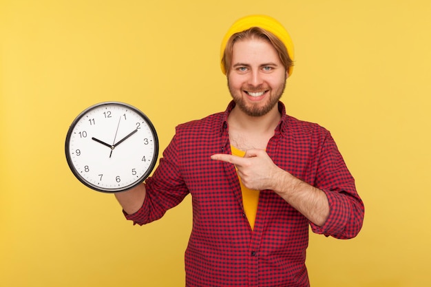 Look at clock, don't rush. Positive hipster guy in checkered shirt pointing big clock and looking at camera with toothy smile, expressing optimism, enough time, not hurry. studio shot isolated