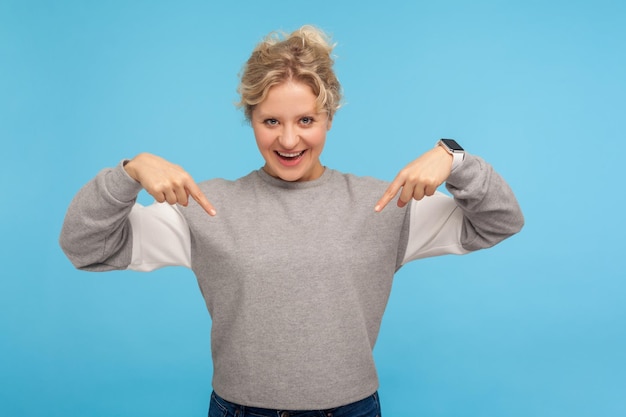 Look down. Happy woman with curly hair in sweatshirt pointing down and smiling happily at camera, showing place below for promotional text, advertising area. studio shot isolated on blue background