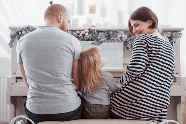 Photo look from behind at mother father and daughter playing white piano