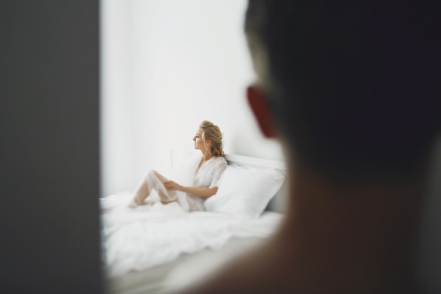 Look from behind naked groom at tender bride sitting on bed