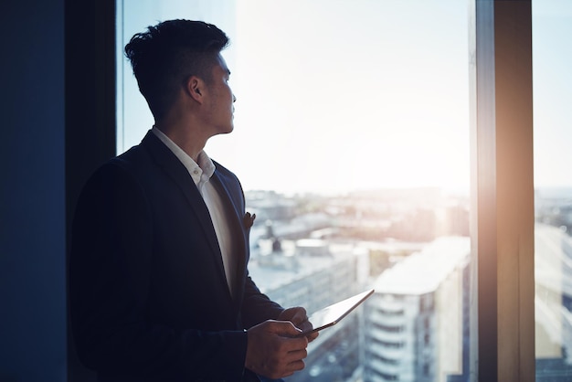 Look to the future of business Shot of a young businessman using a digital tablet while looking out the window in an office