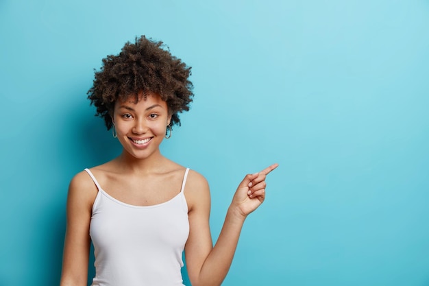 Look here cheerful curly haired young woman indicates right\
shows place for advert or promotional text smiles pleasantly\
dressed in casual t shirt isolated over blue background i have\
idea