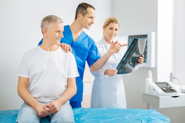 Смотри сюда. Довольный веселый терапевт-мужчина держит пациента за плечо и указывает на рентгеновский снимок, работая со своим коллегой.