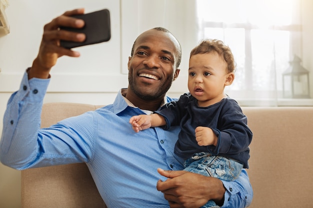이봐. 잘 생긴 쾌활한 젊은 아프리카 계 미국인 아빠가 웃고 셀카를 찍는 동안 작은 곱슬 머리 아들을 들고