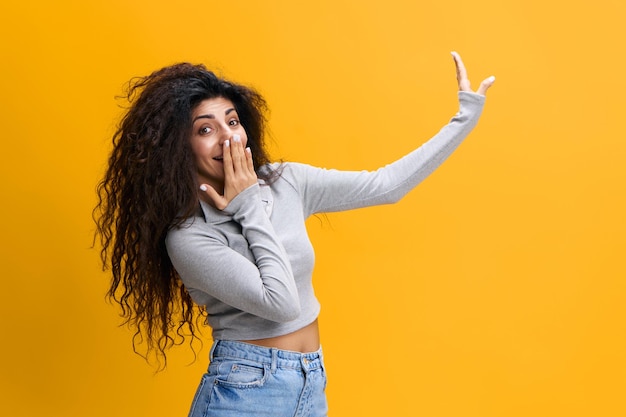 Look here surprised confused young arab lady covers her mouth\
with hand points finger up shows copy space isolated on yellow\
background shocking offer news great sale advertising and app