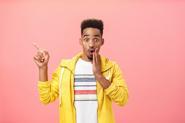 Look I am stunned. Portrait of impressed excited good-looking young male model in stylish yellow jacket opening mouth with gasp covering it with palm pointing amazed at upper left corner