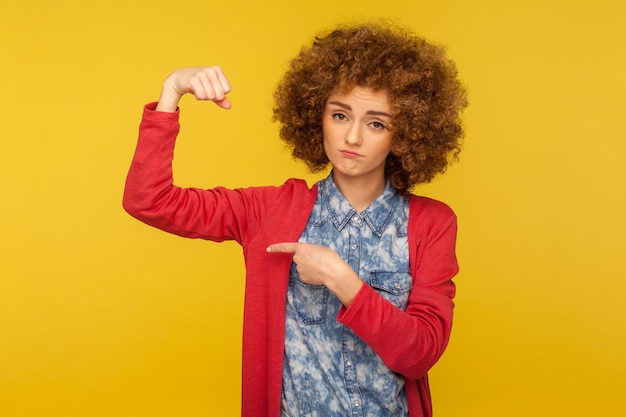 Look I39m strong Portrait of confident woman with curly hair raising arm pointing biceps as metaphor of power and independence female rights concept indoor studio shot isolated on yellow background