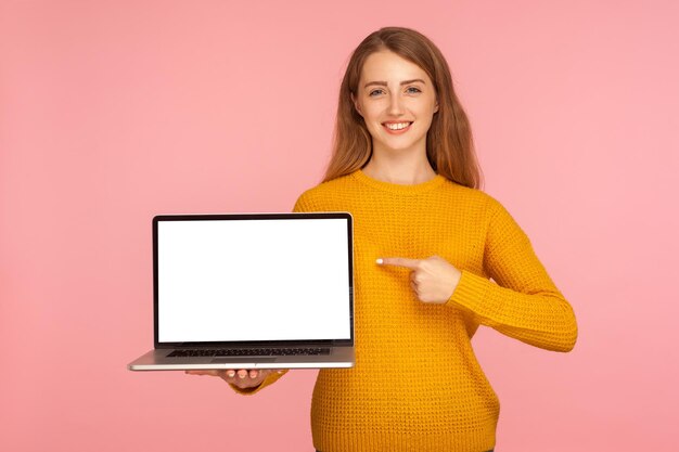 Photo look at internet advertising portrait of cheerful ginger girl in sweater holding laptop and pointing at empty screen copy space for image or text indoor studio shot isolated on pink background