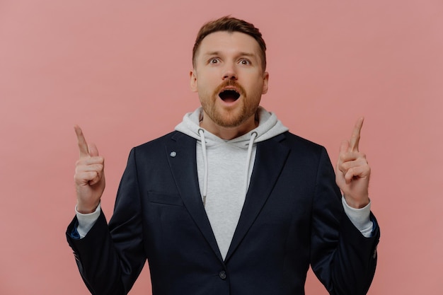 Look its awesome portrait of excited young ginger guy in\
fashionable casual clothes raising index fingers and pointing up\
with amazed face expression promotion and discount concept