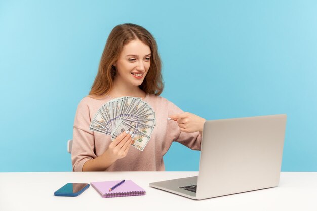 Photo look at money profit. rich happy woman pointing at dollar banknotes while talking on video call on laptop, advertising online eaings, high salary. indoor studio shot isolated on blue background