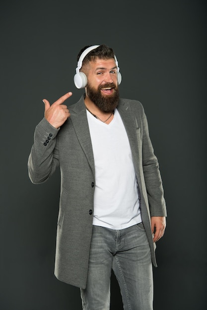 Look at my headset. Hipster point finger at headset grey background. Bearded man listen to music playing in stereo headset. Using wireless bluetooth headset for business and fun. Modern life.