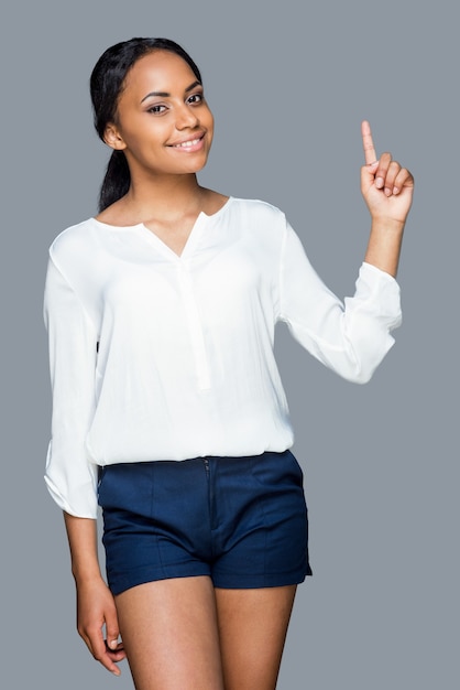 Look at that! Attractive young African woman pointing up and smiling while standing against grey background