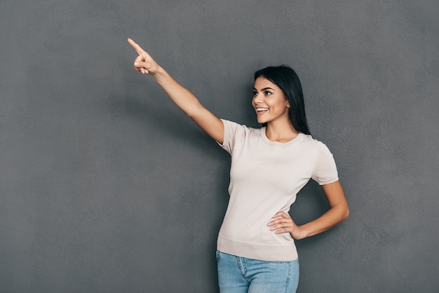 Look over there! Attractive young woman in casual wear pointing away and smiling while standing against grey background