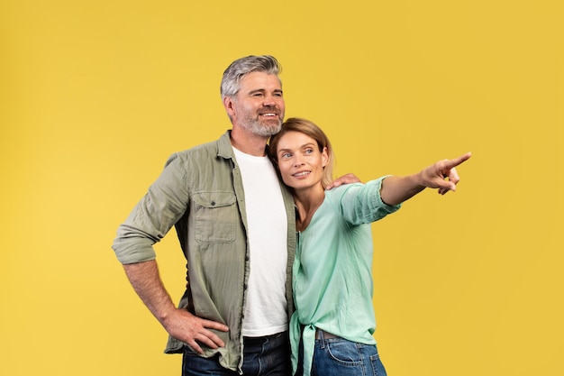 Look there happy mature spouses embracing and woman pointing\
with finger at copy space yellow background