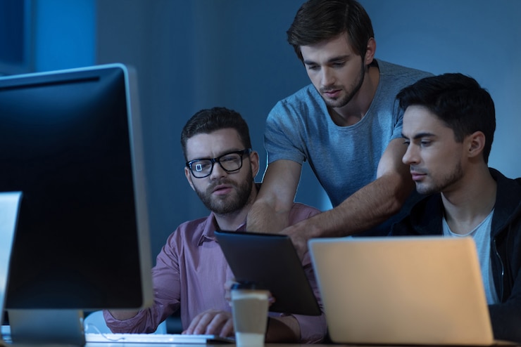 Photo look at this. nice serious attractive man standing behind his colleagues and holding a tablet while showing something to them
