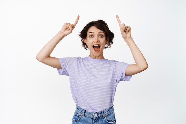 Look up ahead. Smiling happy brunette woman, young girl pointing fingers at top advertisement, promo sale upwards, standing in tshirt against white background.