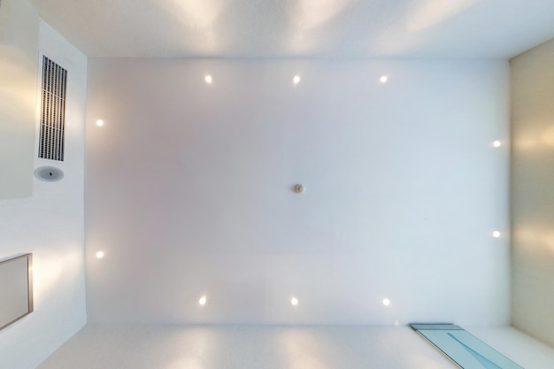 Photo look up on suspended ceiling with halogen spots lamps and drywall construction with fire alarm sensor in empty room in apartment or house stretch ceiling white and complex shape