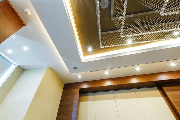 Look up on suspended ceiling with halogen spots lamps and\
drywall construction with fire alarm sensor in empty room in\
apartment or house stretch ceiling white and complex shape