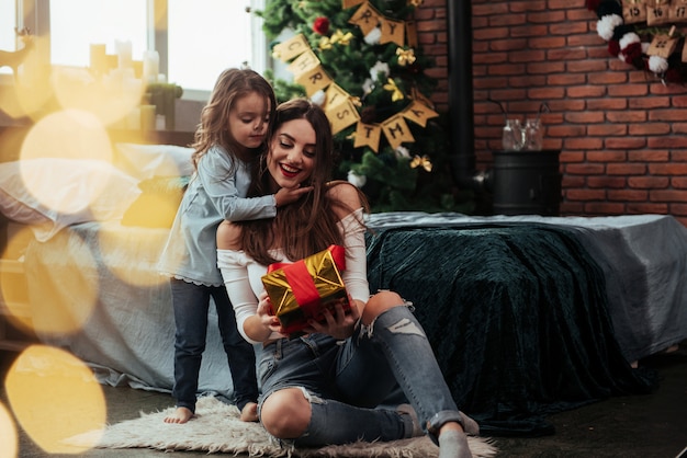 Look what I have for you. Mother and daughter sits in holiday decorated room and holds gift box