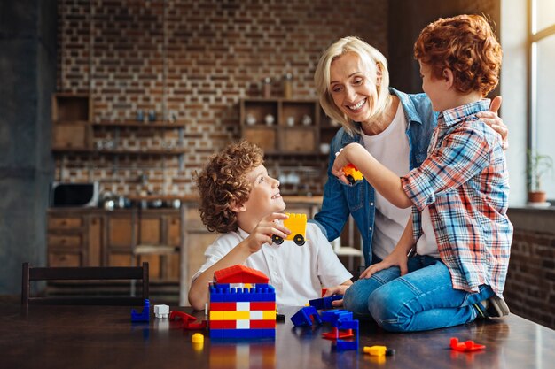 Look what we built. Joyful elderly lady grinning broadly while embracing her grandsons while they playing with a construction set and showing her cars built by themselves.