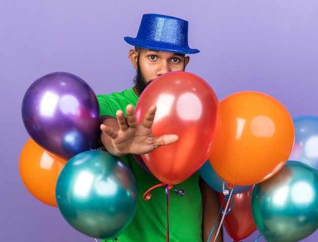 Looking camera young afro-american guy wearing party hat standing behind balloons holding out hand at front isolated on blue wall