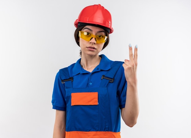 Looking at camera young builder woman in uniform with glasses showing two 