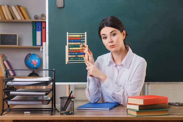 教室で学校の道具を持って机に座ってそろばんを保持しているカメラの若い女性教師を見て