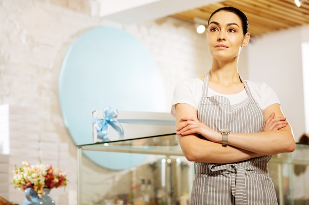 Sembra fiducioso. pasticcere professionista di talento in piedi in un caffè con il braccio incrociato e guardando pensieroso in lontananza