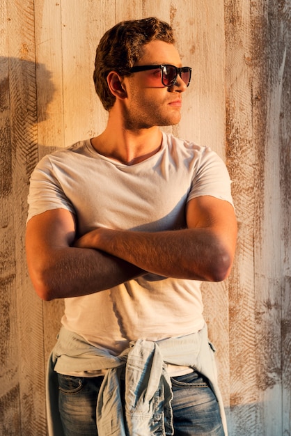 Looking cool. Handsome young man in sunglasses keeping arms crossed while standing against the wooden wall