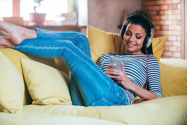 Looking for her favorite song. Beautiful young woman holding mobile phone and looking at it with smile while wearing headphones and sitting on the couch at home