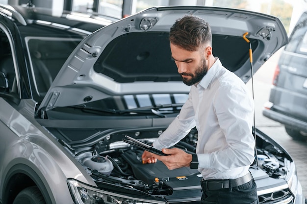 Foto guardando sotto il cofano e tenendo il tablet il giovane in abiti bianchi è nella concessionaria di automobili