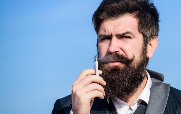 Guardando semplicemente perfetto hipster caucasico brutale con i baffi uomo barbuto che fuma fumo di sigaretta hipster maturo con la barba successo futuro moda formale maschile uomo d'affari contro il cielo