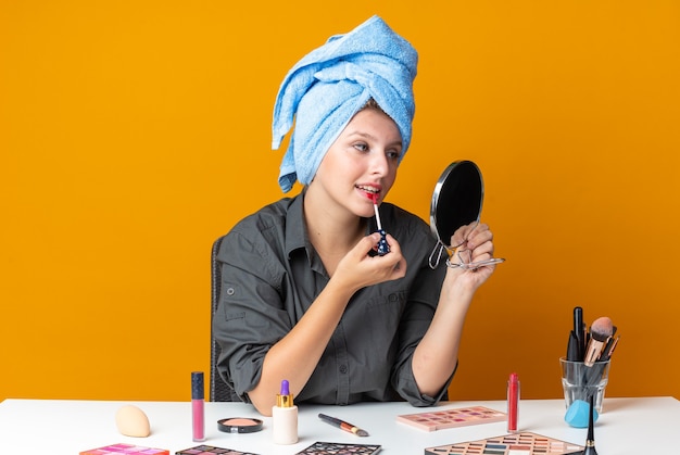 Looking at mirror beautiful woman sits at table with makeup tools wrapped hair in towel applying lip gloss 