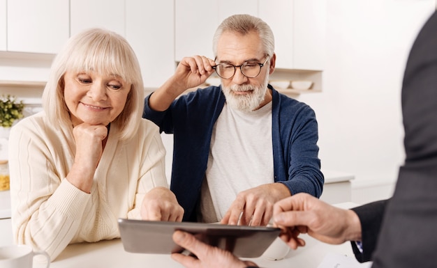 Guardando il nostro futuro progetto di casa. agente immobiliare istruito capace intelligente che lavora con una coppia di clienti in pensione mentre mostra il progetto della casa e utilizza il dispositivo