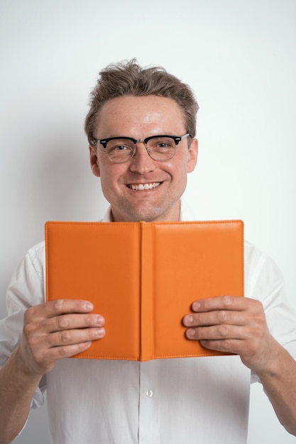 Looking out of orange notebook or journal young man in eye glasses wearing white shirt isolated on white background or in the office Portrait of a shy young businessman holding his diary