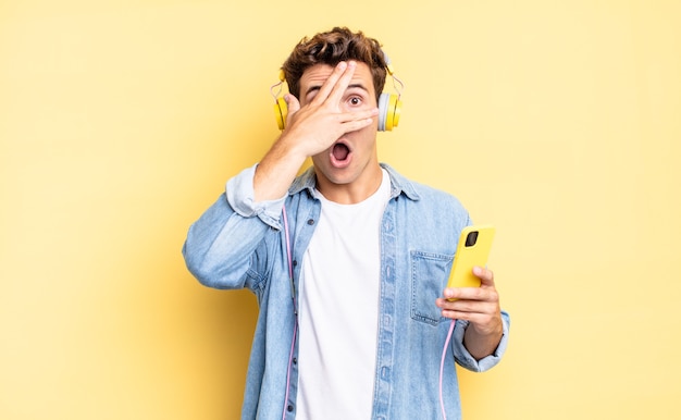 Looking shocked, scared or terrified, covering face with hand and peeking between fingers. headphones and smartphone concept