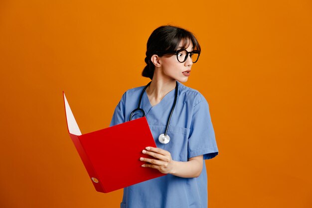 looking at side holding folder young female doctor wearing uniform fith stethoscope isolated on orange background