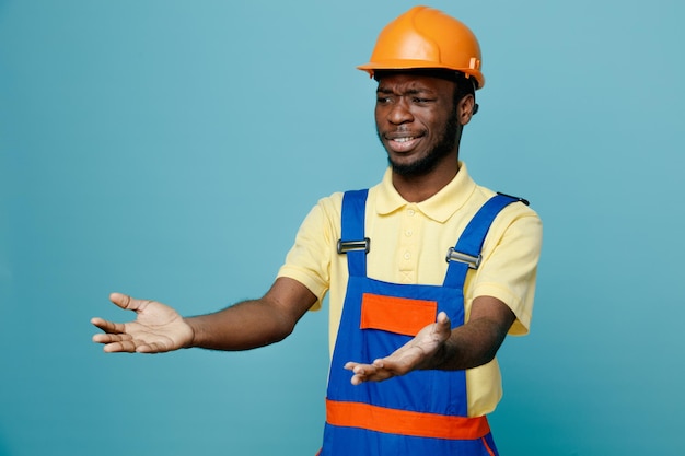 Looking at side holding out hands at side young african american builder in uniform isolated on blue background