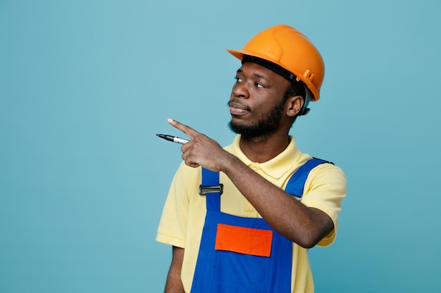 Looking at side points at side young african american builder in uniform holding pencil isolated on blue background