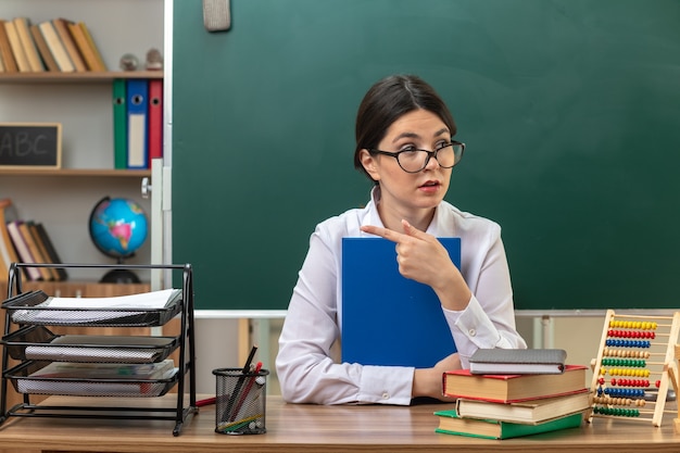 Photo looking side points at side young female teacher wearing glasses holding folder sitting at table with school tools in classroom