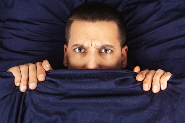 Photo looking straight into the frame a handsome young guy hiding behind a blanket looks to the side direction of gaze in different directions